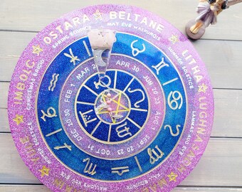 May include: A pink and blue glitter wheel with gold lettering and symbols. The wheel is divided into 12 sections, each representing a month of the year. The wheel also includes the names of the eight pagan holidays, including Ostara, Beltane, Litha, Lughnasadh, Mabon, Samhain, Yule, and Imbolc.