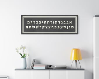 May include: A framed print with Hebrew text in white on a dark background. The print is displayed above a white cabinet with a vase of white flowers, books, and a yellow lamp.