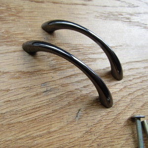 May include: Two black metal cabinet door pulls with a curved design. The pulls are shown on a wooden surface with two screws.