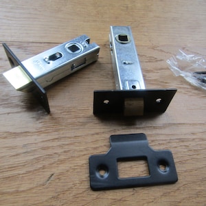 May include: Black door latch set with a brass strike plate and two screws. The latch is made of metal and has a square shape. The strike plate is rectangular and has a small hole in the center. The screws are silver and have a Phillips head.