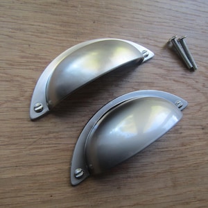 Könnte beinhalten: Zwei silberne Schrankknöpfe mit abgerundeter, leicht gewölbter Form. Die Knöpfe haben eine gebürstete Oberfläche und sind für die Verwendung an Schubladen oder Schränken konzipiert.