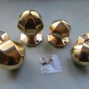 May include: Four brass door knobs with a faceted design. The knobs are in various sizes and are arranged on a light grey surface. Two screws are included in a small plastic bag.