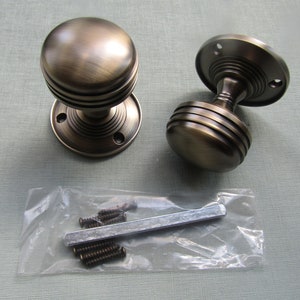 May include: Two antique brass door knobs with a grooved design and a round base. The knobs are shown with screws and a metal spindle in a plastic bag.