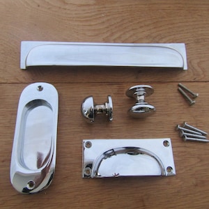 Puede incluir: Un conjunto de componentes de ferretería cromados pulidos, que incluyen un tirador rectangular largo, un tirador empotrado, dos pomos y un tirador rectangular con asa curva. También se incluyen varios tornillos. Los artículos están sobre una superficie de madera.