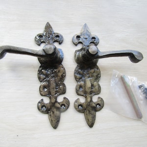 May include: Two black metal door handles with a fleur-de-lis design. The handles have a curved lever and are attached to a square backplate. The handles are shown on a white background with a small bag of screws and a metal rod.
