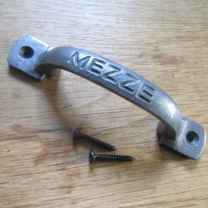 May include: A silver-colored metal cabinet door pull with the word "MEZZE" embossed on the front. The pull comes with two black screws.