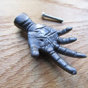 May include: A cast iron hand-shaped door knob or drawer pull in a dark grey finish. The knob has five fingers and a screw is included for installation.