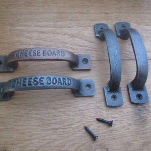 May include: Two sets of black metal handles with the words "Cheese Board" engraved on them. The handles are on a wooden surface with two screws.