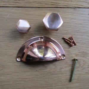 May include: Three copper cabinet knobs and a copper cup-shaped drawer pull. The knobs are hexagonal and the pull is curved. The knobs and pull are on a wooden surface.