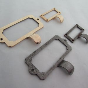 以下が含まれることがあります： さまざまな仕上げの金属製ラベルホルダーが4つ。2つは長方形で、湾曲したハンドルが付いており、1つは金色のトーン、もう1つはより暗く、テクスチャ加工された仕上げです。2つの小さなラベルホルダーも表示されています。