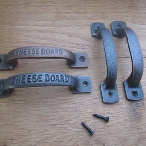 May include: Four cast iron cheese board handles in various shades of gray and bronze. Two handles are inscribed with the words "CHEESE BOARD". Two black screws are also visible.