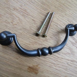 Puede incluir: Un tirador de cajón de metal negro con un mango redondeado y una perilla decorativa. El tirador está fijado a una superficie de madera con dos tornillos.