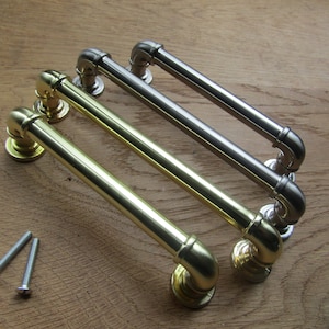 Peut inclure: Trois poignées de porte de style industriel en finition or, argent et nickel. Les poignées sont en métal et ont un design de tuyau. Les poignées sont présentées sur une surface en bois.