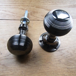 Può includere: Due pomelli per porte in nichel nero con un design a coste. I pomelli sono rotondi e hanno una base piatta.