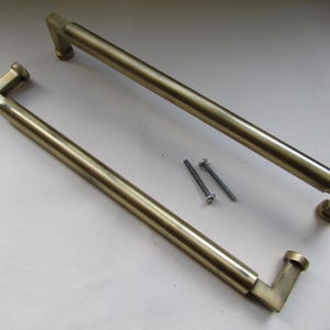 Könnte beinhalten: Zwei messingfarbene Schrankgriffe mit rechteckigem Design sowie zwei silberne Schrauben. Die Griffe haben eine röhrenförmige Mitte und quadratische Enden. Die Hardware ist für Möbel und Heimdekoration konzipiert.