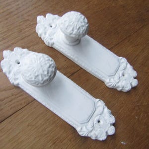 May include: Two white, ornate door knobs with a decorative floral design. The knobs are made of metal and have a smooth, polished finish.