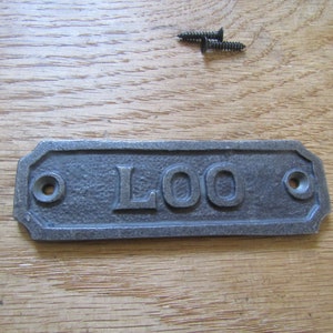 May include: A grey cast iron sign with the word "LOO" in raised letters. The sign has two holes for mounting.