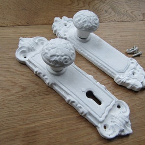 Può includere: Due pomelli per porte bianchi decorativi con buchi per le chiavi e dettagli decorativi. I pomelli sono su una superficie di legno.