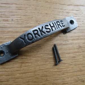 Puede incluir: Un tirador de puerta de hierro fundido plateado con la palabra "YORKSHIRE" en relieve en la parte superior. El tirador viene con dos tornillos.