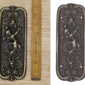 May include: An antique bronze door plate with a detailed relief sculpture of a cherub and floral designs. The rectangular plate is about 15 cm long, with intricate detailing and a vintage appearance. Includes two screw holes for mounting.