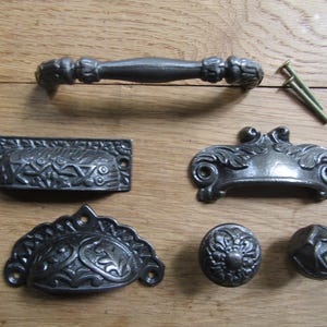 May include: Set of six black cast iron drawer pulls with ornate designs. The pulls include a long handle, two small pulls with a curved shape, and three round knobs.