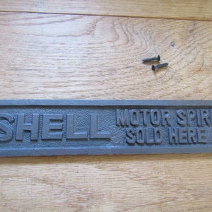 Może przedstawiać: Czarny znak z żeliwa z napisem "SHELL MOTOR SPIRIT SOLD HERE" w wypukłych literach.