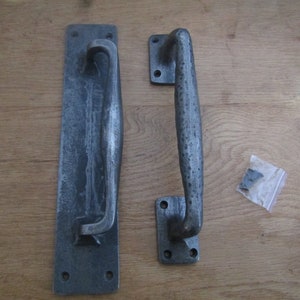 May include: Two antique-style door handles in a dark grey finish. The handles are made of metal and have a simple, elegant design. The handles are shown on a wooden surface.