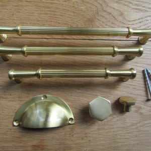 以下が含まれることがあります： 真鍮製のキャビネット金具のコレクション。3つのリブ付きバープル、半月型のプル、六角形のノブ、小さな丸いノブが含まれています。2本のネジも含まれています。木製の表面に金具が配置されています。