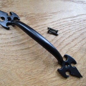 Könnte beinhalten: Schwarzer Metalltürgriff mit spitzem, dekorativem Design. Der Griff hat einen gebogenen Griff und ist mit Schrauben an einer Holzoberfläche befestigt.