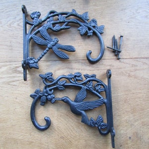 Może przedstawiać: Dwa czarne metalowe wsporniki do roślin z motywem ważki i kolibra. Każdy wspornik ma haczyk na dole.