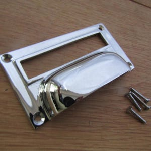 Könnte beinhalten: Ein glänzender, verchromter Schubladengriff mit rechteckiger Grundplatte und gebogenem Griff. Enthält vier Schrauben zur Montage. Die Hardware ist für Schränke, Schubladen oder andere Möbel konzipiert.