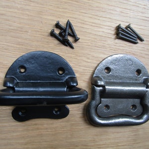 Könnte beinhalten: Zwei Metallkoffergriffe, einer schwarz und einer grau, mit passenden Schrauben. Die Griffe haben eine halbkreisförmige Oberseite mit einem scharnierartigen, abgerundeten Griff. Die Schrauben sind dunkel und scheinen zur Befestigung der Griffe auf einer Holzoberfläche zu dienen.