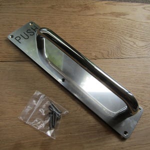 May include: A silver door handle with a rectangular plate that says "PUSH" in black lettering. The handle is attached to a wooden surface.