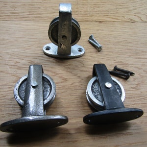 Könnte beinhalten: Drei Metallrollen mit silberner Oberfläche und eine schwarze Rolle. Die Rollen sind auf einer flachen Basis mit zwei Schraubenlöchern montiert. Die Rollen werden zum Aufhängen von Gegenständen oder zum Bewegen von Dingen verwendet.