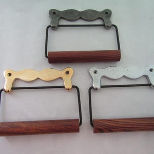 May include: Three toilet paper holders with different metal finishes: gold, silver, and a darker gray. Each has a dark wood dowel for the toilet paper roll and a black metal frame. The holders are displayed on a white surface.