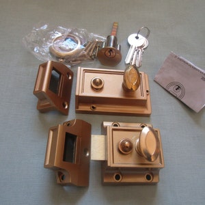 May include: A set of two gold-toned door locks with a key and screws. The locks are square shaped with a round knob on the side. The key is silver and has a small loop on the end. The screws are small and silver.