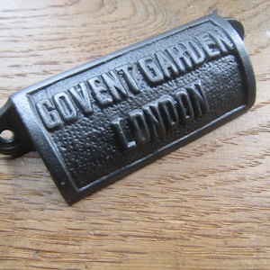 Könnte beinhalten: Ein schwarzes, rechteckiges Metallschild mit dem geprägten Text "COVENT GARDEN LONDON". Das Schild hat eine strukturierte Oberfläche und einen gebogenen Griff mit einem Befestigungsloch. Es liegt auf einem Holzgrund.