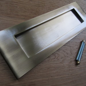 Pode incluir: Uma placa de correio de níquel escovado com uma abertura retangular. A placa é projetada para instalação em uma porta ou parede. Inclui dois parafusos para instalação.