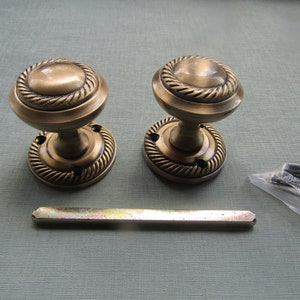 Peut inclure: Deux poignées de porte antiques en laiton avec un motif de corde autour de la poignée circulaire. L'image montre également une barre métallique et un petit sachet de vis. La quincaillerie a une esthétique vintage.
