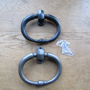 May include: Two door knockers, one black and one silver, with a rounded oval shape and a decorative knob on the bottom. The knockers are on a wooden surface.