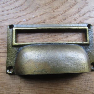 以下が含まれることがあります： 長方形のラベルホルダーと湾曲したハンドルが付いた、アンティーク真鍮の引き出しプル。 金具は風化した仕上げで、取り付け用のネジ穴が4つあります。 プルは木製の表面に置かれています。