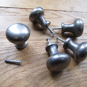 May include: Five silver metal cabinet knobs with a rounded, slightly domed shape. The knobs are arranged in a circle on a wooden surface. One screw is lying on the surface.