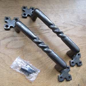 Może przedstawiać: Dwa czarne żeliwne klamki do drzwi z zakręconym wzorem i kwadratowymi płytkami. Klamki mają około 15 cm długości. Klamki są pokazane na drewnianej powierzchni z dwoma śrubami w plastikowym woreczku.