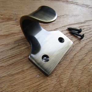 May include: A bronze-colored metal hook with a curved design and a diamond-shaped base. Two black screws are included. The hook is designed for hanging items and is suitable for various home decor styles.