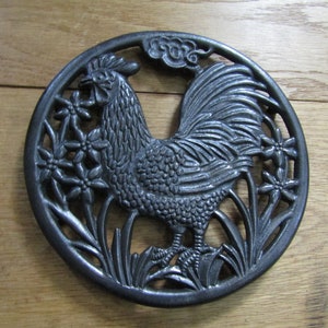May include: A black cast iron trivet featuring a rooster design with flowers and leaves. The rooster is standing on a bed of leaves and flowers.