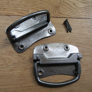 May include: Two metal handles with a rustic finish. The handles have a rectangular shape with a rounded edge and a metal loop for pulling. The handles are designed for attaching to a box or drawer.