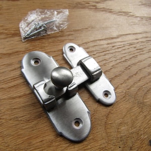 May include: Silver-toned metal latch with a rounded knob. The latch has two rectangular plates with rounded edges and a decorative design. The latch is attached to a wooden surface.