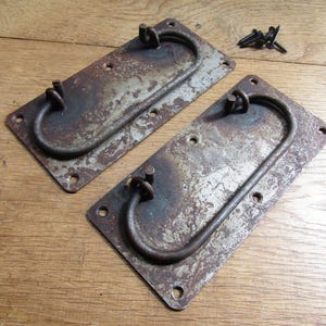 Könnte beinhalten: Zwei antike Metall-Schubladengriffe mit einem verwitterten, rostigen Finish. Jeder Griff hat eine rechteckige Grundplatte mit einem gebogenen Griff und Befestigungsmaterial. Ein kleiner Haufen Schrauben ist sichtbar.