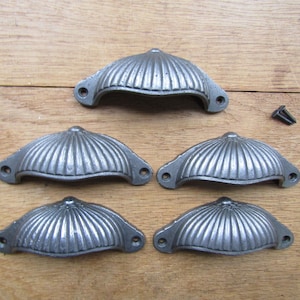 May include: Five silver-toned cast iron cabinet drawer pulls with a scalloped design. The pulls are arranged in a row on a wooden surface.