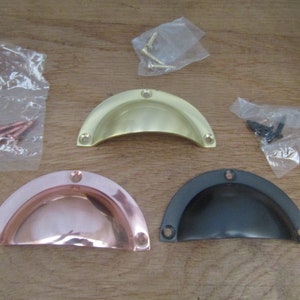 Könnte beinhalten: Drei Metall-Muschelgriffe in Messing, Kupfer und Dunkelgrau, mit passenden Schrauben in kleinen Plastiktüten. Die Griffe sind halbkreisförmig und haben Schraubenlöcher.